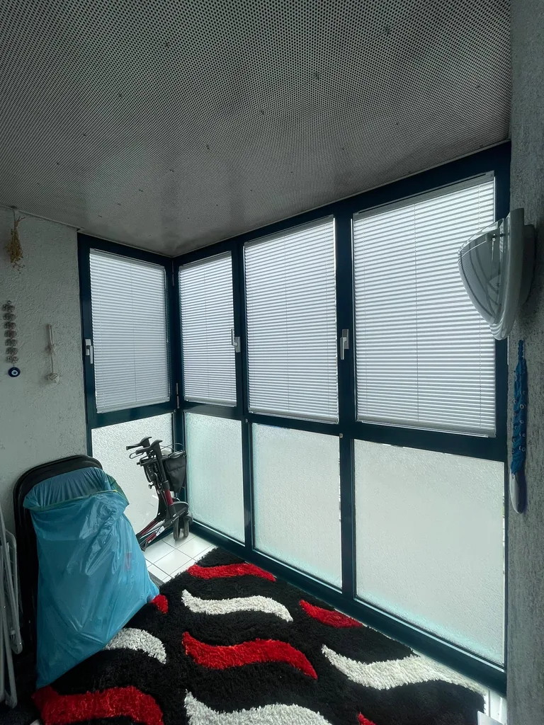 Geschlossene weiße Plissees an der dunklen Fensterfront eines verglasten Balkons